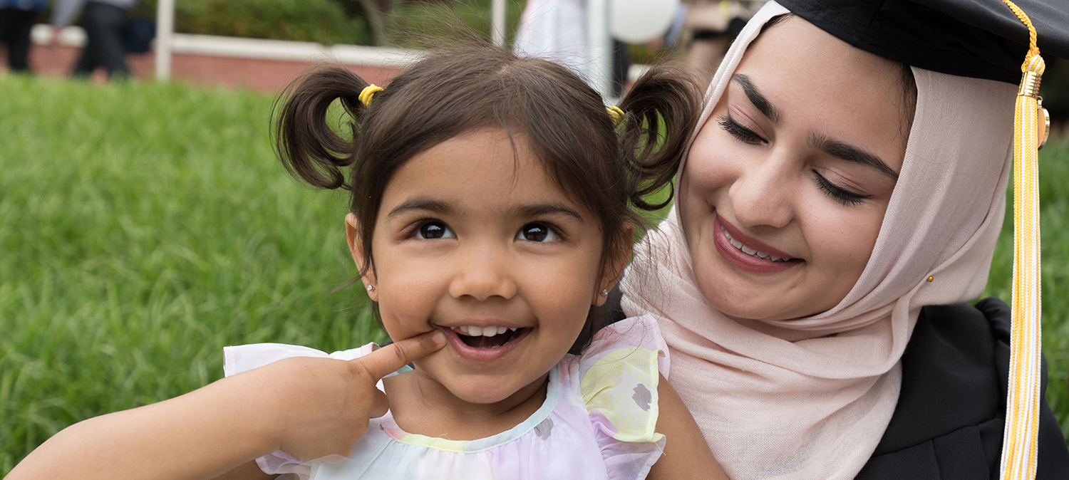 A female child smiling. Her mother in cap and gown looks at her smiling too.