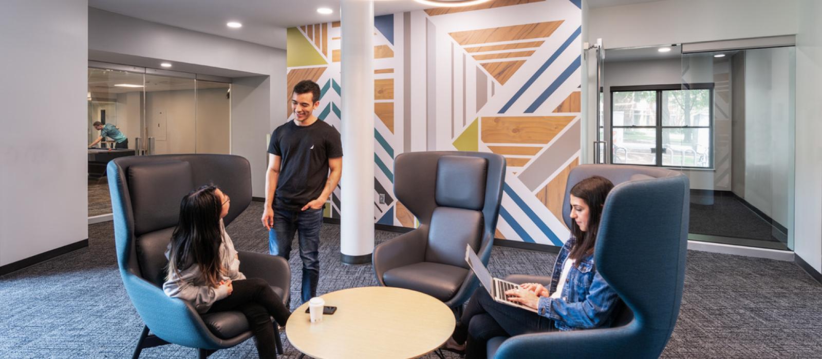 A young woman and a young man speak at the lobby while another young woman writes on her laptop computer.