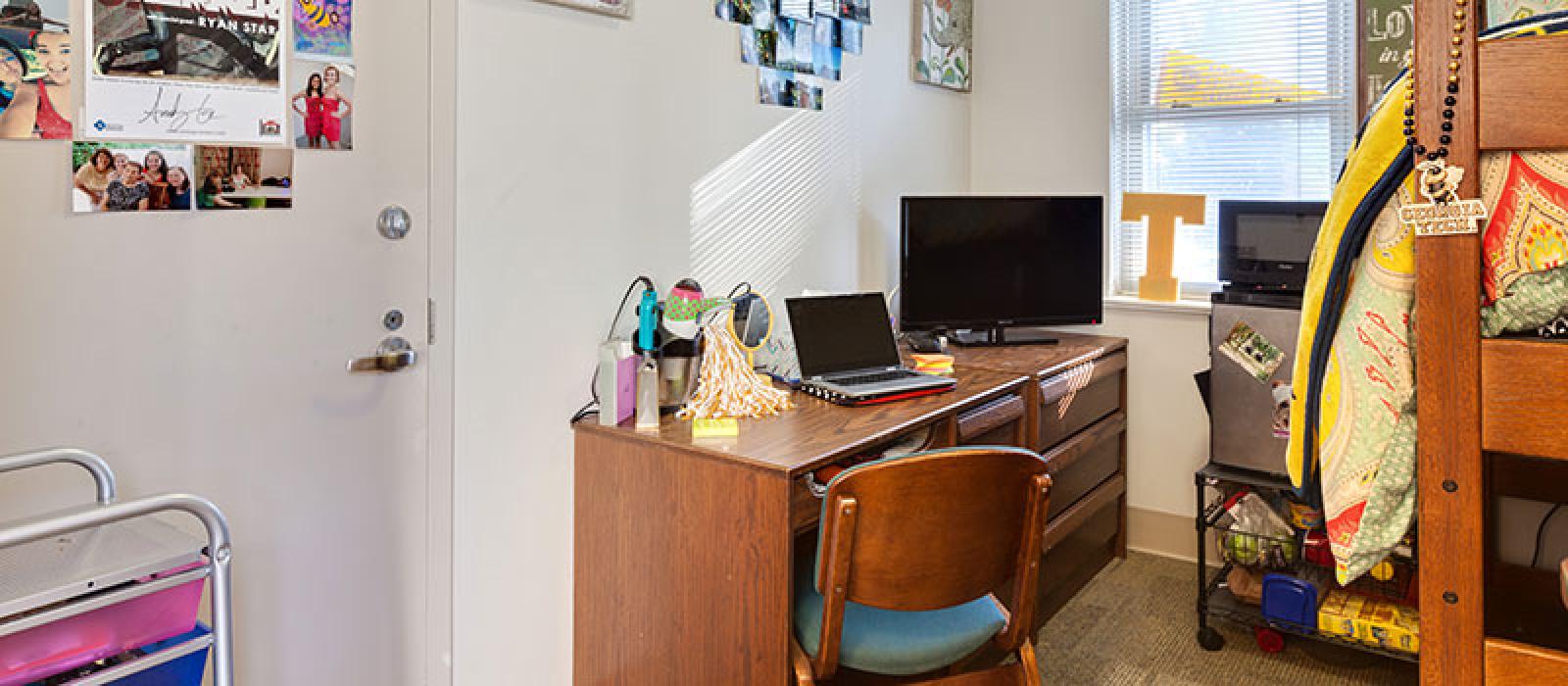 The corner of a bunk bed, a desk with a laptop and TV monitor on top and a desk chair.