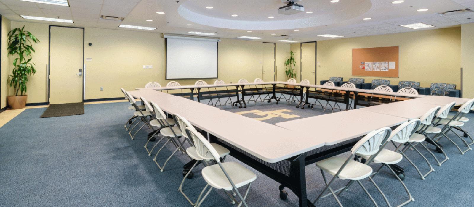 Activity room with long tables in the center in a square formation. Chairs around it, a wall screen, the GT logo in the center.