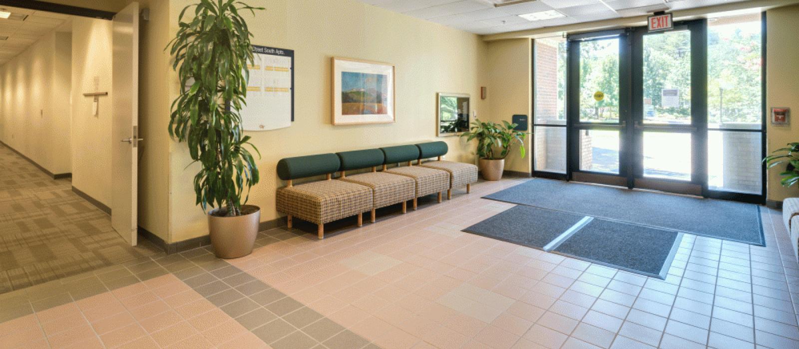 Atrium. Big glass doors, four chairs together forming a type of sofa. Two plants on each side, one taller than the other.