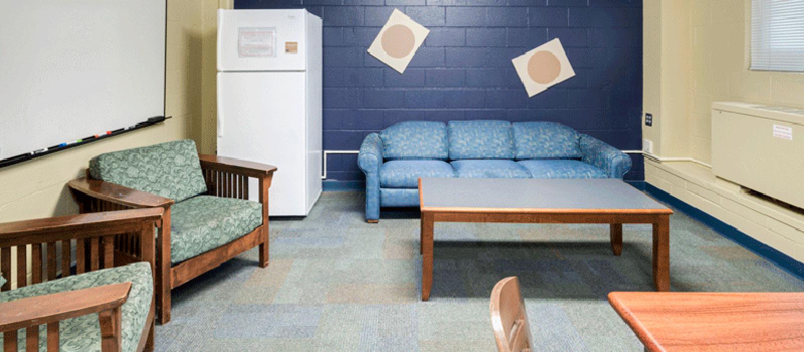 Lounge with a sofa, a refrigerator, 2 arm chairs and the corner of a table and chair.