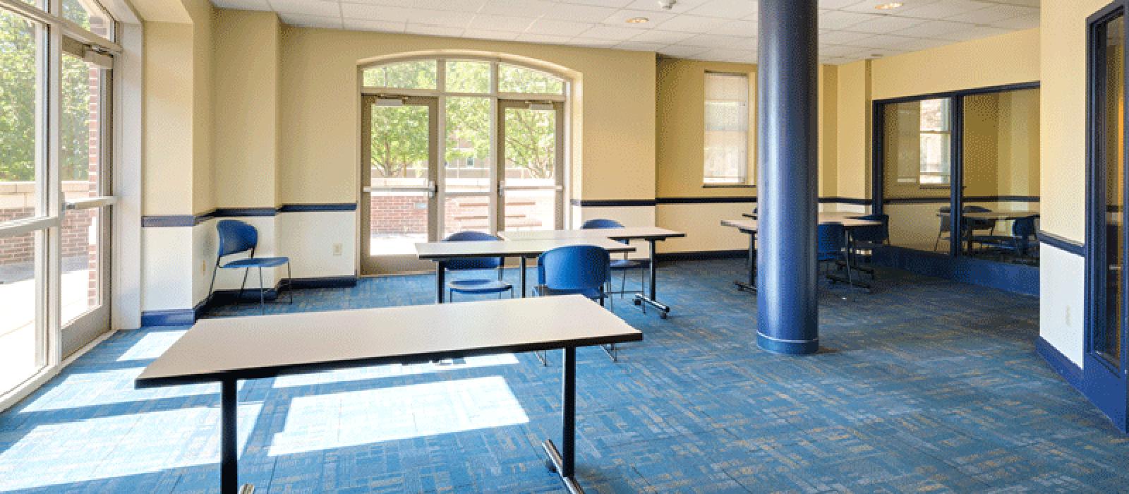 Sunny activity room with big glass doors, long and square tables and chairs.